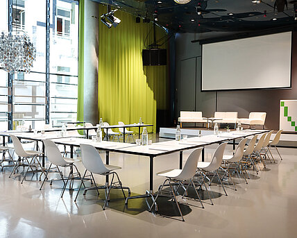 Im Space04 im Kunsthaus Graz stehen weiße Tische mit Sesseln vor dem Podium im Hintergrund. Die Leinwand für Projektionen ist heruntergelassen. Auf dem Podium stehen weiße Couchen sowie ein Rednerpult.